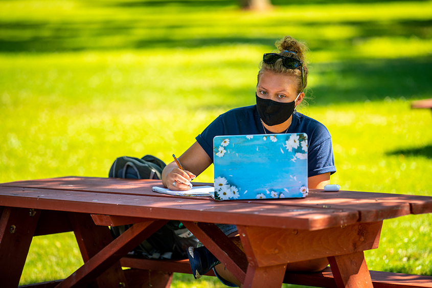 Student sitting at a picnic table outside writing on paper and using a laptop.