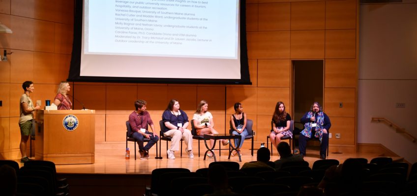 Six students sit on stage with two moderators standing by podium
