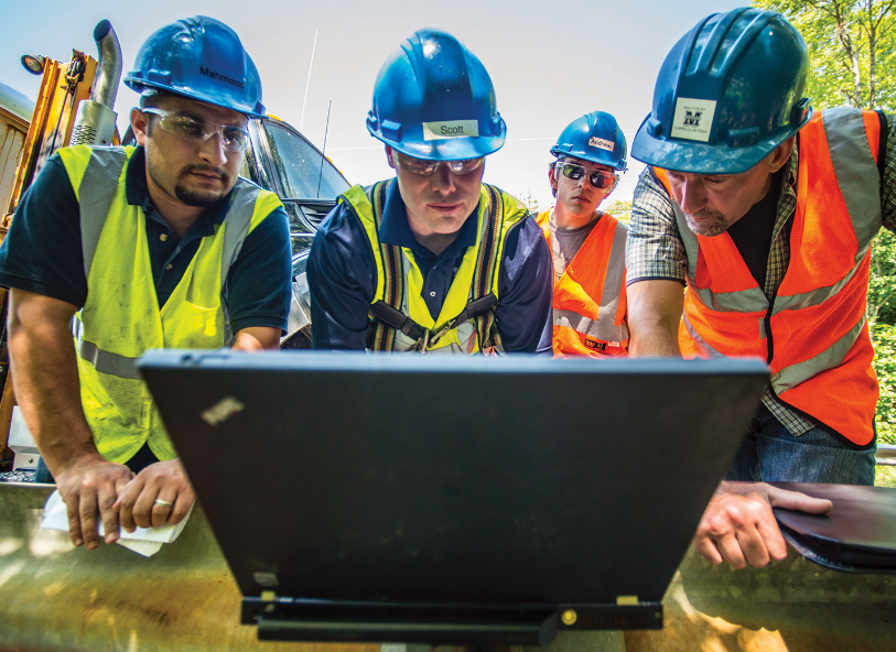 a group of engineers in hard hats and safety vests looking at laptop computer